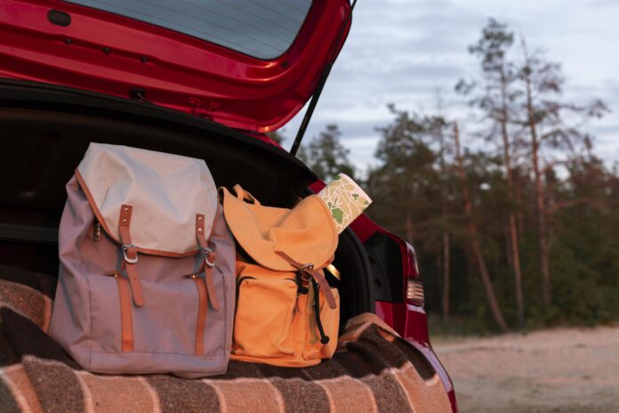 Mochilas no porta-malas de um carro vermelho, com um mapa e uma paisagem arborizada ao fundo, capturando o momento de preparação para uma viagem.