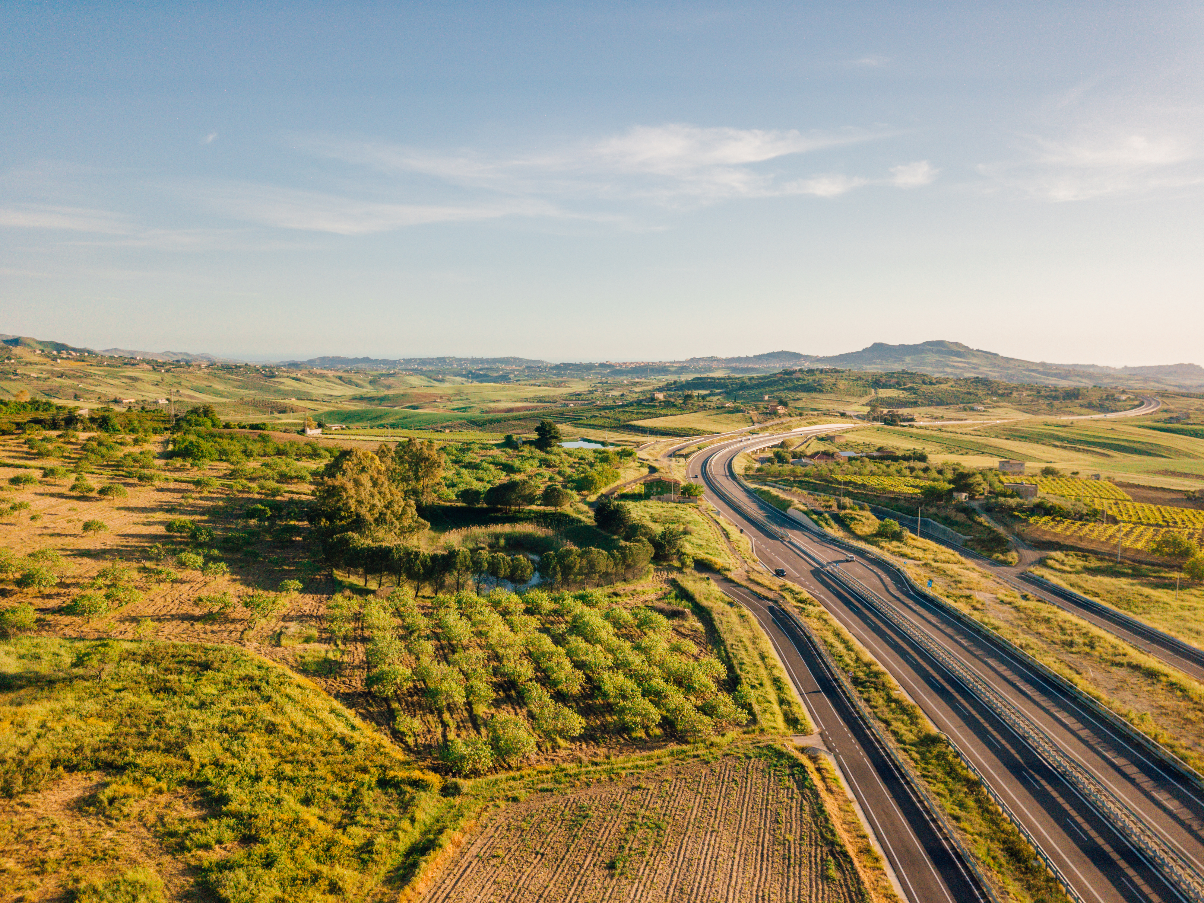 Estrada moderna cercada por campos verdes e plantações em um cenário montanhoso, capturada ao pôr do sol.