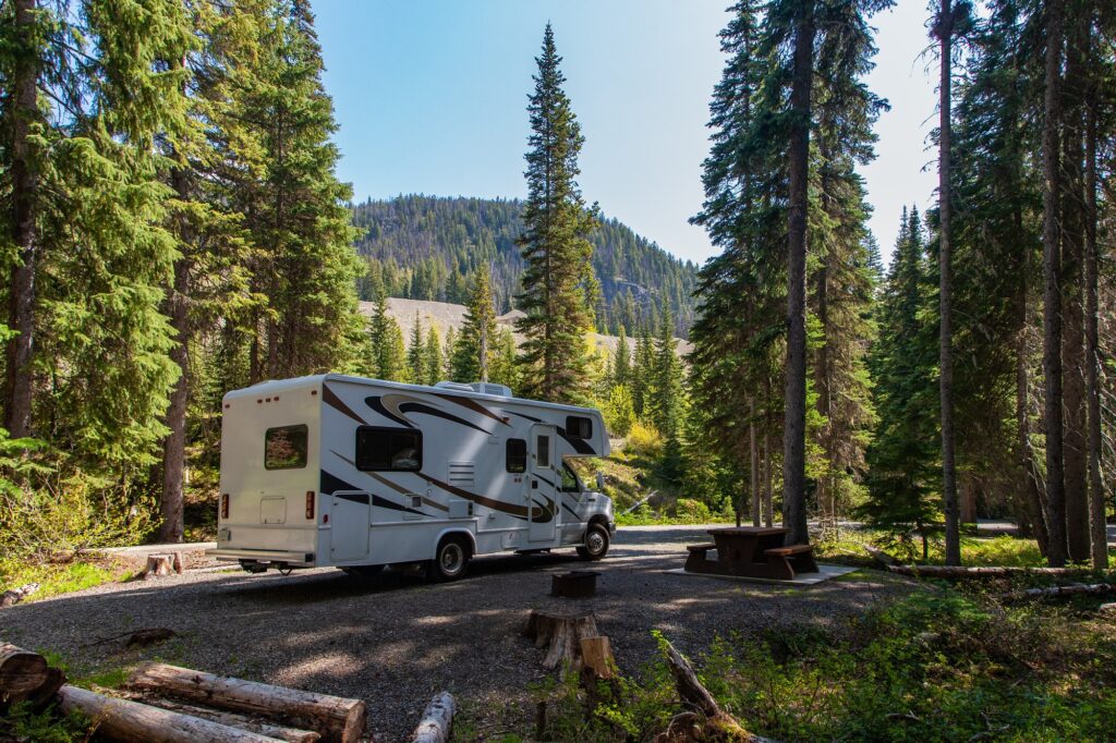 Motorhome branco estacionado em meio a uma floresta com árvores altas e montanhas ao fundo, destacando viagens na natureza.