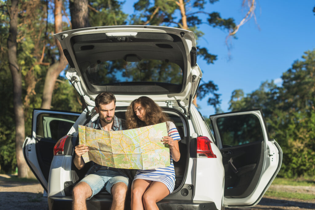 Casal sentado no porta-malas de um carro branco, analisando um mapa de papel em meio a um cenário arborizado e ensolarado.